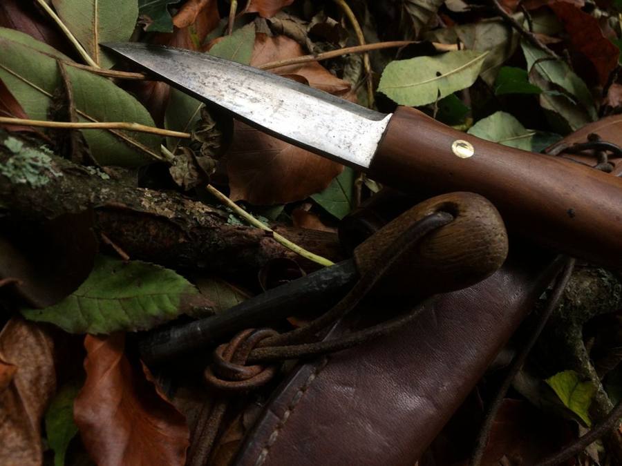 A wooden-handled knife and fire steel resting on a leather sheath among beech leaves
